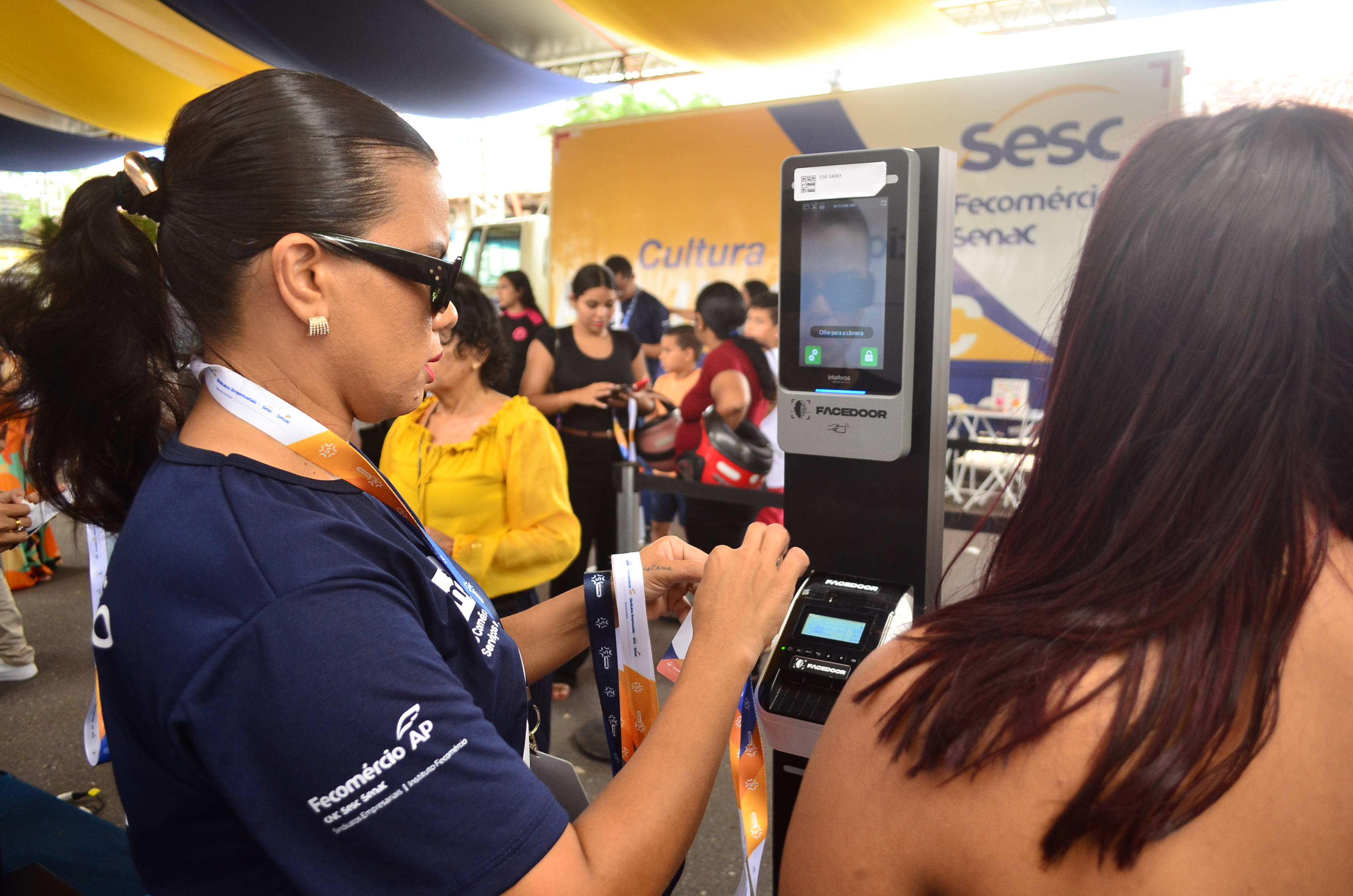 Atendente junto ao equipamento da Facedoor, que exibe Olhe para a câmera durante o credenciamento