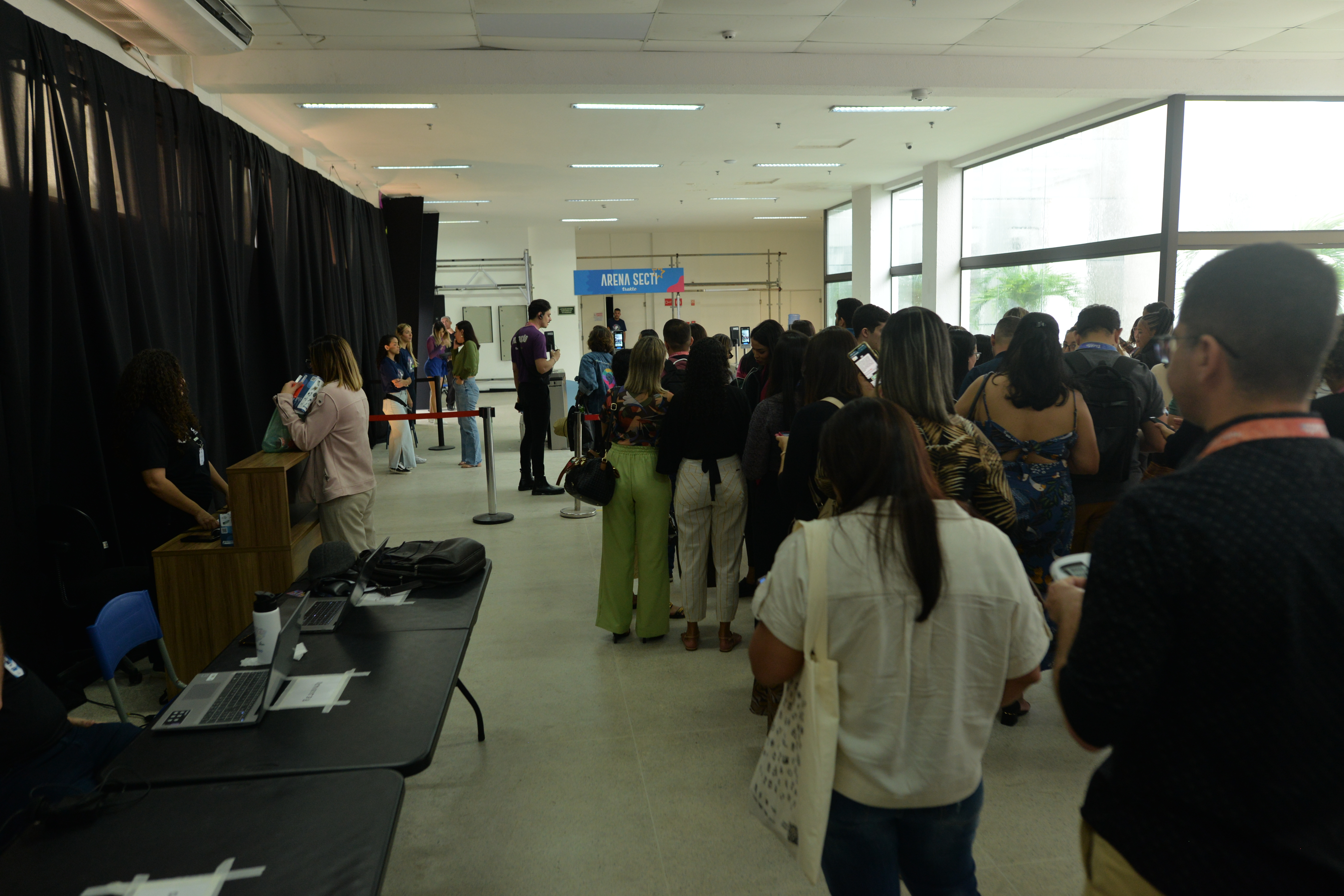 Fila de participantes no hall do evento, com os equipamentos de acesso ao fundo e a equipe de credenciamento em atendimento